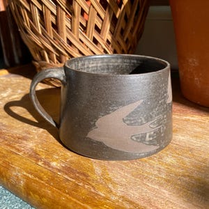 Puede incluir: Taza de cerámica marrón oscuro con asa y diseño de pájaro marrón claro. La taza está colocada sobre una superficie de madera. Una cesta de mimbre y una maceta de terracota son visibles en el fondo. Ideal para café.