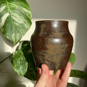 Könnte beinhalten: Eine kleine, handgefertigte braune Keramikvase mit breitem Boden und leicht ausgestelltem Rand. Die Vase hat eine strukturierte Oberfläche mit Farbvariationen, die von Dunkelbraun bis zu helleren Beigetönen reichen. Die Vase wird in einer Hand gehalten, mit einem grünen und weißen Blatt im Hintergrund.
