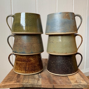 May include: Six handmade ceramic mugs stacked on a wooden surface. The mugs are in shades of green, blue, brown, and black. Each mug has a handle and a flared shape. The mugs are made of ceramic.