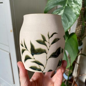 May include: A cylindrical ceramic vase with a matte off-white, textured surface. The exterior is decorated with a hand-painted botanical pattern featuring dark green, glossy leaves and thin stems that wrap around the body of the vessel.