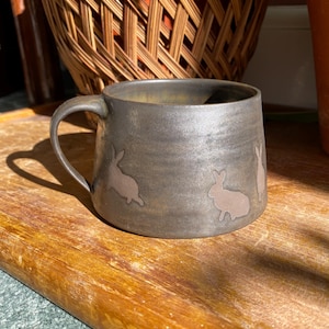 Puede incluir: Una taza de cerámica hecha a mano con un esmalte gris oscuro y un asa curva. La taza presenta tres siluetas de conejo marrón claro alrededor del exterior. La taza está sobre una superficie de madera.