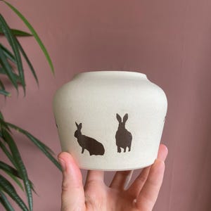 Puede incluir: Un jarrón de cerámica de color crema con forma redondeada. El jarrón presenta siluetas marrones oscuras de conejos alrededor del exterior. El jarrón está sostenido en la mano de una persona, con un fondo rosa y una planta verde visible.