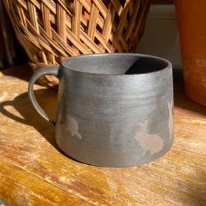 Puede incluir: Una taza de cerámica gris oscuro con asa, con un patrón repetido de siluetas de conejos marrones claros. La taza está sobre una superficie de madera, con una cesta tejida y una maceta de terracota en el fondo.