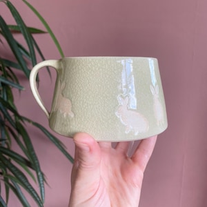Puede incluir: Una taza de cerámica verde claro con un esmalte craquelado y un asa curva. La taza presenta tres ilustraciones de conejos blancos. La taza está sostenida en la mano de una persona.