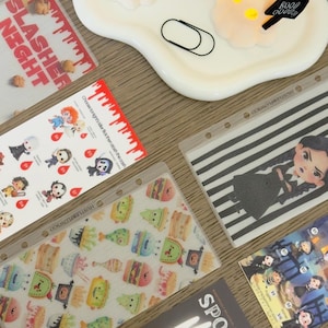 以下が含まれることがあります： ハロウィーンをテーマにしたステッカー、メニュー、装飾品のコレクション。ステッカーには漫画のキャラクターや不気味なお菓子が描かれています。「SPOOKY MENU」というテキストが入ったメニュー。白いトレイには、幽霊の形をした装飾品が置かれています。