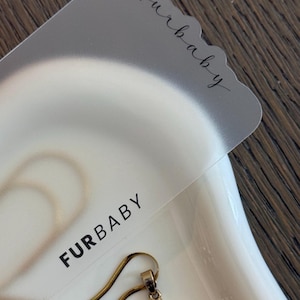 May include: A translucent frosted display card featuring the text "furbaby" in a cursive script and "FURBABY" in a bold sans-serif font. A gold-toned chain necklace with a crucifix pendant rests beneath the card on a smooth white surface.