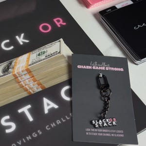 Könnte beinhalten: Ein schwarzer Schlüsselanhänger mit einer silbernen Kette und einem schwarzen Anhänger, auf dem "Slack or Stack" steht. Der Anhänger ist an einer schwarzen Karte befestigt, auf der "Hustle and Flow, Chain Game Strong" und "Lock this in your binder & stay locked in to stack your savings, no slacking!" steht.