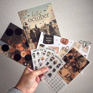 Puede incluir: Una colección de rastreadores de ahorro y hojas de pegatinas con temática de Halloween que presentan ilustraciones de calabazas, fantasmas y brujas. Los artículos incluyen textos como hello october, hustle hunny, monster mash, costume party y hocus pocus.