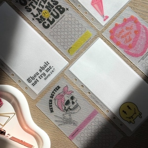 May include: A selection of clear, rectangular planner inserts with printed designs and text. One insert displays "OVERSTIMULATED MOMS CLUB" with a smiley face. Another reads "Thou shalt not try me. mom 24:7". Other inserts feature a skull and a cake.