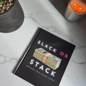 May include: A black book with the title "Slack or Stack Savings Challenge Book" in white text. A stack of cash is pictured on the cover.