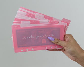 May include: Pink plastic binder with clear pockets and tabs labeled with months and events, such as "Vacation #2", "Money Tree", and "52 Stay at Home". The binder has a clear pocket on the front that shows a dollar bill with the text "create your luck" printed on it.