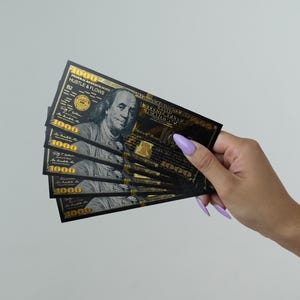 May include: A stack of black $1000 bills with gold accents and the words "Hustle & Flows" printed on them. The bills are fanned out in a person's hand.
