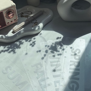May include: A collection of clear, rectangular planner inserts with the seasons: Summer, Fall, Winter, and Spring. A white tray holds jewelry and a pen, with a small radio in the background. The items are on a white surface.