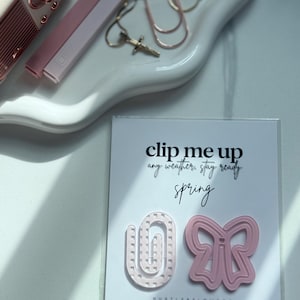 May include: A white tray holds pink and gold office supplies, including paper clips and a small radio. A card displays two pink paper clips, one shaped like a bow and the other a staple, with the text "clip me up any weather, stay ready spring."