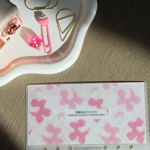 May include: A white ceramic tray with a pink rim holds gold paper clips, a pink paper clip with a strawberry design, and a pink die. A clear plastic card with a pink balloon dog pattern and text that reads "SMALLSHOPS" and "HUSTLE&FLOWSCO" is also present.