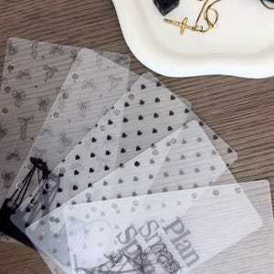 Può includere: Un set di cinque divisori per agenda rettangolari e traslucidi con fori pre-perforati sul lato. I design includono disegni a linee nere di fiocchi, piccoli cuori e un'illustrazione floreale con il testo stampato Plan, Spend, Growth.