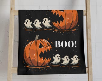 Könnte beinhalten: Schwarzes Halloween-Handtuch mit zwei orangefarbenen Kürbissen mit geschnitzten Gesichtern und dem Wort "BOO!" in Weiß. Mehrere weiße Geisterfiguren sind ebenfalls abgebildet. Das Handtuch hängt an einem Holzregal.