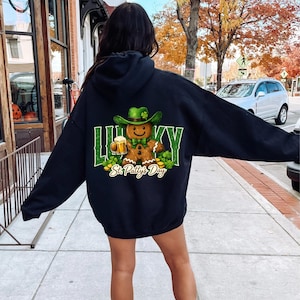Może przedstawiać: Czarna bluza z kapturem z grafiką na Dzień Świętego Patryka. Grafika przedstawia piernikowego ludzika w zielonym kapeluszu trzymającego piwo, ze słowem "Lucky" i "St. Patty's Day" w kolorze zielonym.