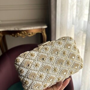 May include: White clutch bag with detailed gold and pearl decoration. The rectangular design features a geometric pattern of gold beads, small mirrors, and pearl accents. A gold clasp secures the top. A stylish accessory for special occasions.