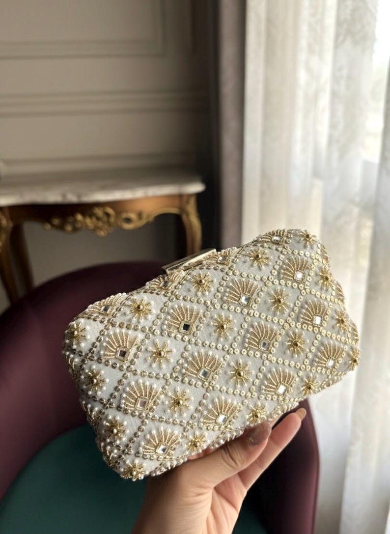 May include: White clutch bag with detailed gold and pearl decoration. The rectangular design features a geometric pattern of gold beads, small mirrors, and pearl accents. A gold clasp secures the top. A stylish accessory for special occasions.