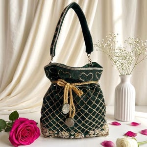 Pode incluir: Uma bolsa de veludo verde escuro com bordados dourados em padrão de losangos. A bolsa tem um fecho de cordão dourado e uma alça a condizer. Uma rosa rosa e flores brancas ao fundo.