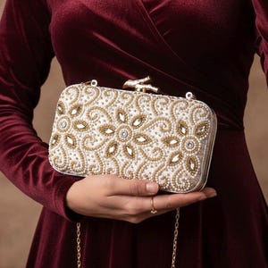 May include: A rectangular, off-white clutch with intricate beadwork in gold and clear crystals. The clutch features a floral design and a gold chain strap. The clasp is gold, and the bag is held against a burgundy velvet dress.