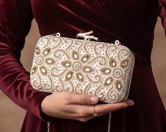 May include: A rectangular, off-white clutch with intricate beadwork in gold and clear crystals. The clutch features a floral design and a gold chain strap. The clasp is gold, and the bag is held against a burgundy velvet dress.