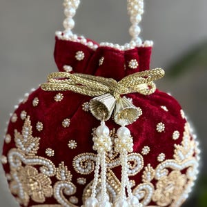 Może przedstawiać: Okrągła, czerwona aksamitna torebka z misternymi złotymi i perłowymi koralikami. Torebka posiada perłowy uchwyt i frędzel oraz złote zapięcie na kokardę. Projekt zawiera kwiatowe wzory i małe perłowe akcenty.
