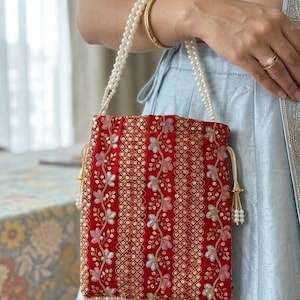 May include: A red embroidered drawstring bag with a pearl beaded handle and tassel fringe. The bag features floral and geometric patterns in gold, pink, and silver. The background is a light blue garment.