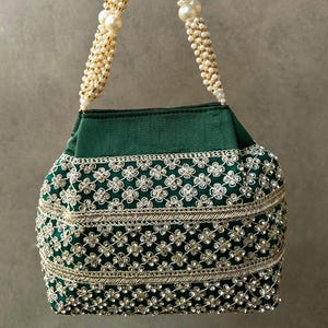 Könnte beinhalten: Eine grüne Handtasche mit einem Perlenhenkel und aufwendiger floraler Stickerei. Die Tasche hat eine dunkelgrüne Stoffbasis, verziert mit goldenen und silberfarbenen Details. Der Henkel besteht aus kleinen weißen Perlen und größeren Perlen.