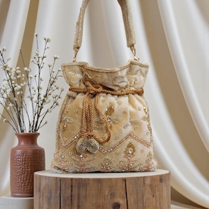 May include: A beige velvet handbag with intricate gold embroidery and beadwork. The bag features a drawstring closure and a matching gold cord handle. The design includes floral and geometric patterns, creating an elegant and ornate accessory.