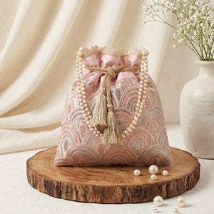 Puede incluir: Un bolso rosa con cordón, asa de cuentas de perlas y borlas. El bolso presenta un diseño bordado y se exhibe sobre una rodaja de madera con perlas dispersas. Un jarrón con flores blancas se encuentra en el fondo.
