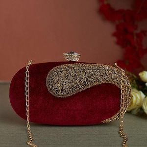Peut inclure: Une pochette en velours bordeaux avec une chaîne dorée et un grand ornement en strass. L'ornement est en forme de motif paisley.