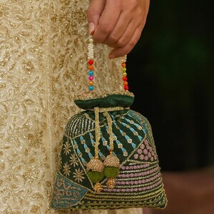 Könnte beinhalten: Ein dunkelgrüner Beutel mit Kordelzug und aufwendigen Stickmustern in Türkis, Rosa und Gold. Die Tasche hat einen Perlenhenkel mit bunten Perlen und goldenen Quasten. Die Tasche wird von einer Person gehalten, die ein goldbesticktes Kleidungsstück trägt.