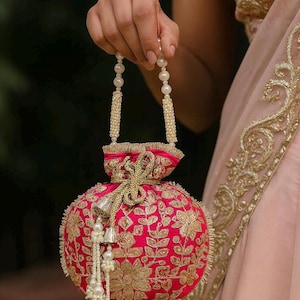 Puede incluir: Un bolso de mano bordado en rosa vibrante con patrones florales dorados. El bolso presenta un asa de cuentas de perlas y detalles de borlas. El diseño es ornamentado y elegante, adecuado para ocasiones especiales.