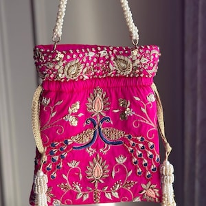May include: A pink fabric pouch with a pearl chain strap and gold and blue embroidery of peacocks. The pouch has a drawstring closure and tassels.