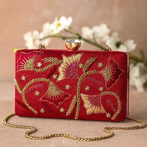 May include: A rectangular red clutch with gold floral embroidery and a gold chain strap. The clutch features a gold clasp with a clear gem. The floral design includes gold sequins and small gold stars.