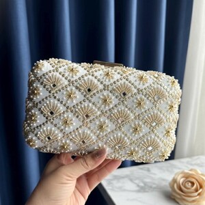 May include: White beaded clutch bag with gold and pearl embellishments. The rectangular bag features a geometric pattern of gold accents, small pearls, and square mirrored details. A gold clasp secures the top.