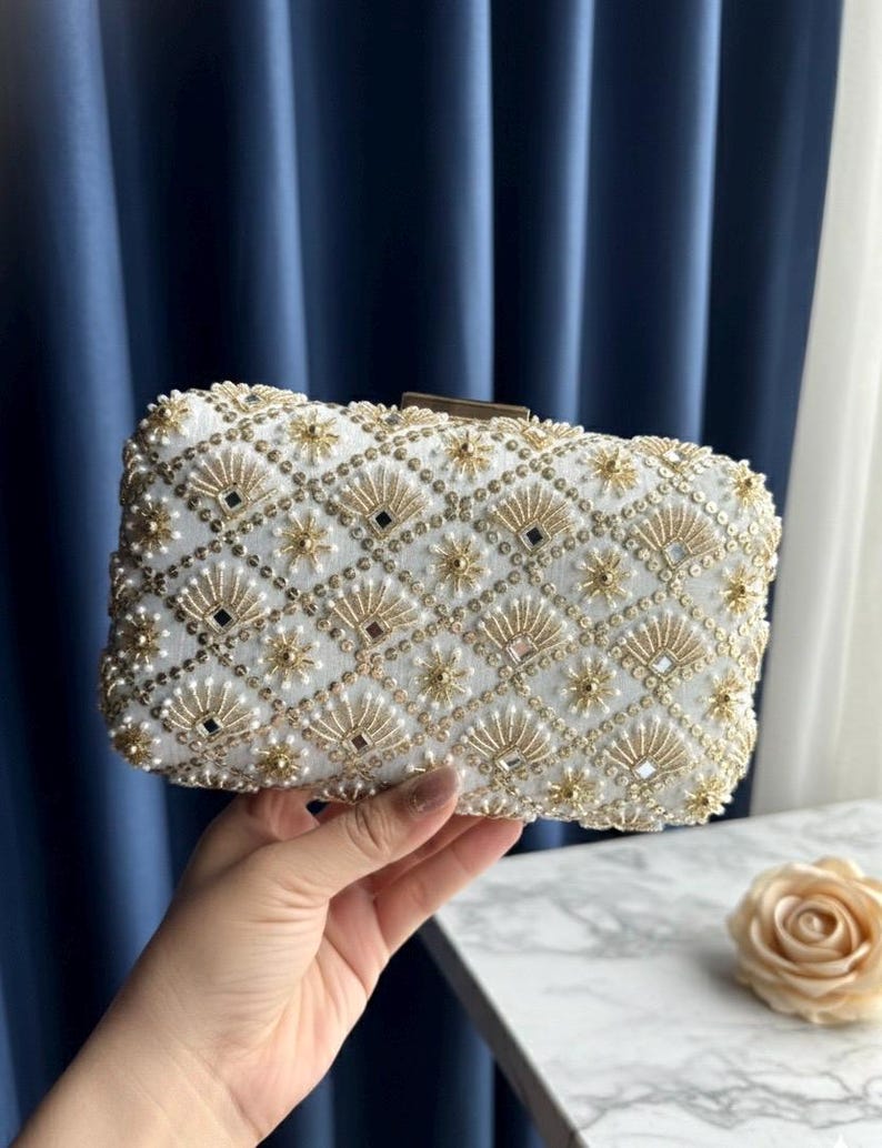 May include: White beaded clutch bag with gold and pearl embellishments. The rectangular bag features a geometric pattern of gold accents, small pearls, and square mirrored details. A gold clasp secures the top.