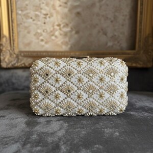 May include: A white beaded clutch bag with a geometric pattern of gold beads and small pearls. The bag has a rectangular shape and a clasp closure. The background is a blurred image of a framed painting.