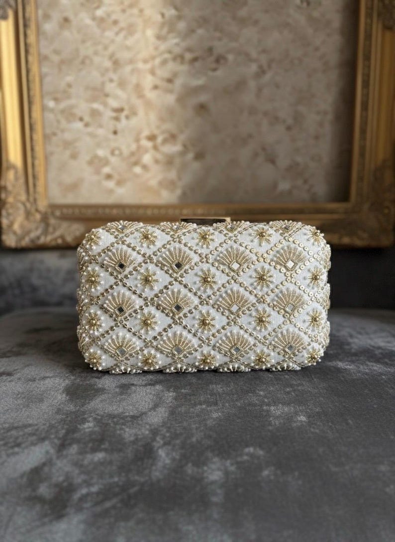 May include: A white beaded clutch bag with a geometric pattern of gold beads and small pearls. The bag has a rectangular shape and a clasp closure. The background is a blurred image of a framed painting.