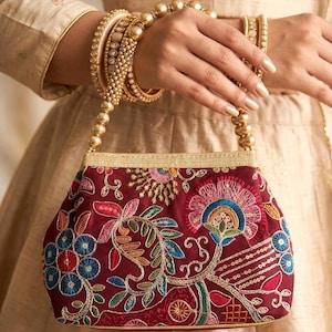 May include: A burgundy handbag with intricate floral embroidery in various colors. The bag features a gold beaded handle and trim. The image also shows gold bangles and a matching outfit, suggesting a formal or festive occasion.