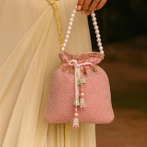 May include: A pink beaded pouch bag with a pearl handle. The bag has a drawstring closure with gold trim and decorative tassels. The bag is held by a person.