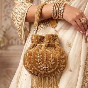 May include: A mustard-colored velvet handbag with intricate gold embroidery and a beaded fringe. The bag features a drawstring closure and a gold chain strap. The image also shows gold bangles, a ring, and a cream-colored garment with gold embellishments.