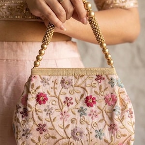 Puede incluir: Un bolso rosa rubor con bordado floral en tonos rojos, rosas y azules. El bolso presenta asas y ribetes con cuentas doradas. El diseño floral está detallado con hilo dorado y lentejuelas.