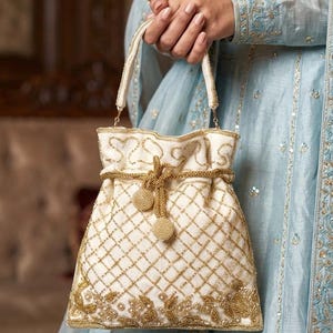 May include: Ivory drawstring handbag with gold embroidery. The bag features a diamond pattern and floral accents. The handles and drawstring are also adorned with gold detailing. The bag is held against a light blue garment.