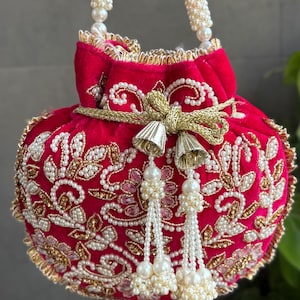 May include: A vibrant fuchsia velvet handbag with intricate gold and pearl embellishments. The bag features a beaded handle and decorative tassels. The design includes floral patterns and a gold bow, creating an elegant accessory.