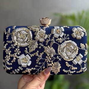 May include: Navy blue rectangular clutch bag with detailed gold and silver floral embroidery. The bag has a decorative clasp and is a stylish accessory for special occasions.