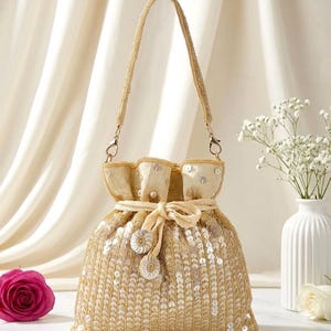 May include: A gold drawstring bag with a beaded handle and a sequined body. The bag has a cream-coloured top with pearl accents and a decorative bow with floral embellishments. The bag is designed for carrying small items.