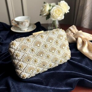 May include: Ivory rectangular clutch bag with gold and pearl detailing. The beaded design has a geometric pattern with fan and starburst shapes. A gold clasp secures the top. The bag sits on a dark blue velvet surface.